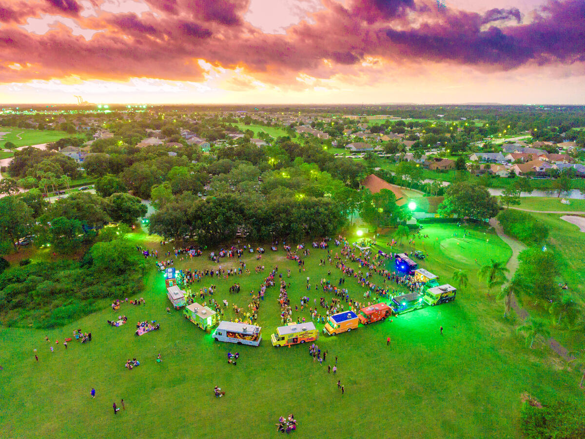Aerial view of a Florida food truck rally at dusk, trucks ringed by string lights and a crowd on the grass